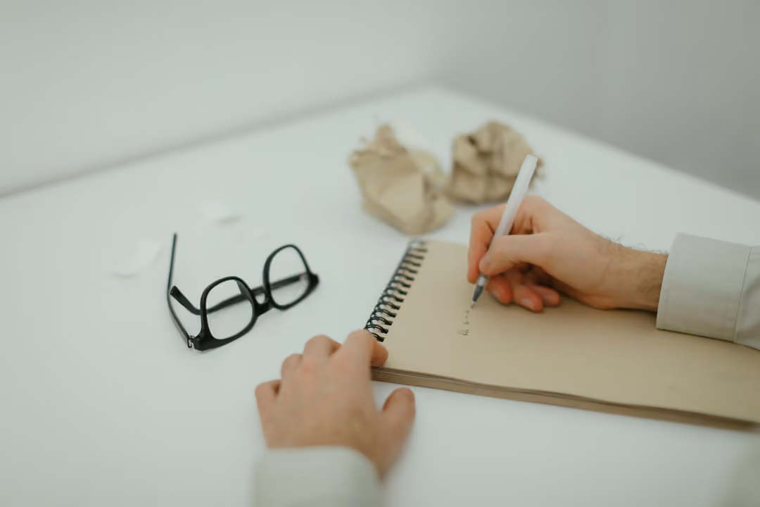 A candidate drafts notes by hand in a notebook at a clean desk, glasses and crossed-out drafts nearby.