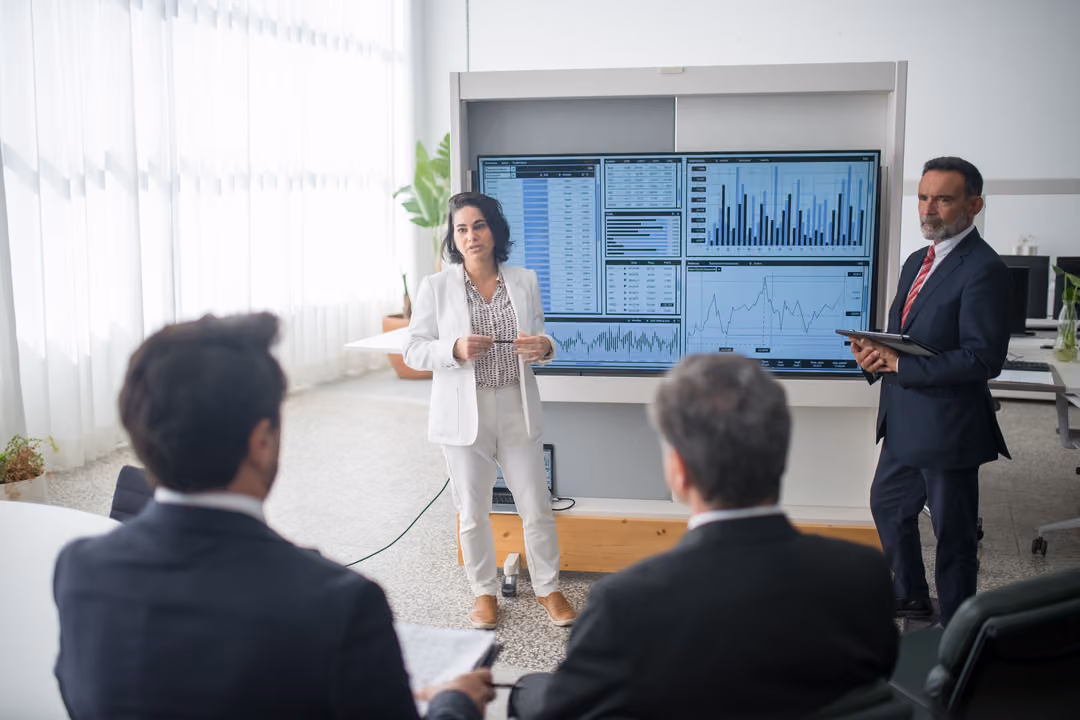 An analyst presents a data dashboard on a wall-mounted screen to colleagues seated in a bright meeting room.