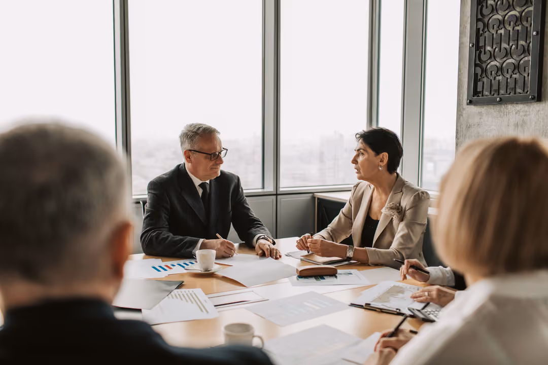 Two senior executives in conversation at a boardroom table overlooking the city, papers and notes between them.