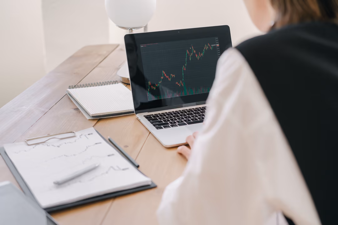 A market chart on a laptop screen beside a hand-drawn price sketch on a notepad, at a wooden desk.